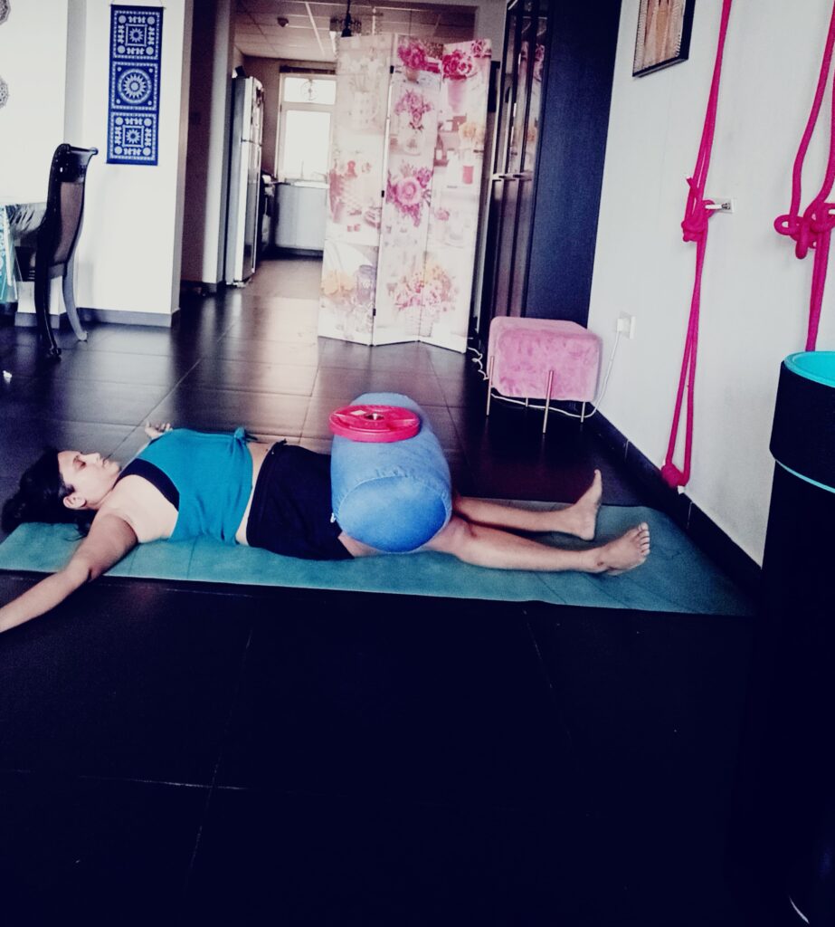 "A woman practicing Savasana, the corpse pose, with a blue bolster and a red plate weight on top for added pressure, lying on a yoga mat in a well-equipped studio